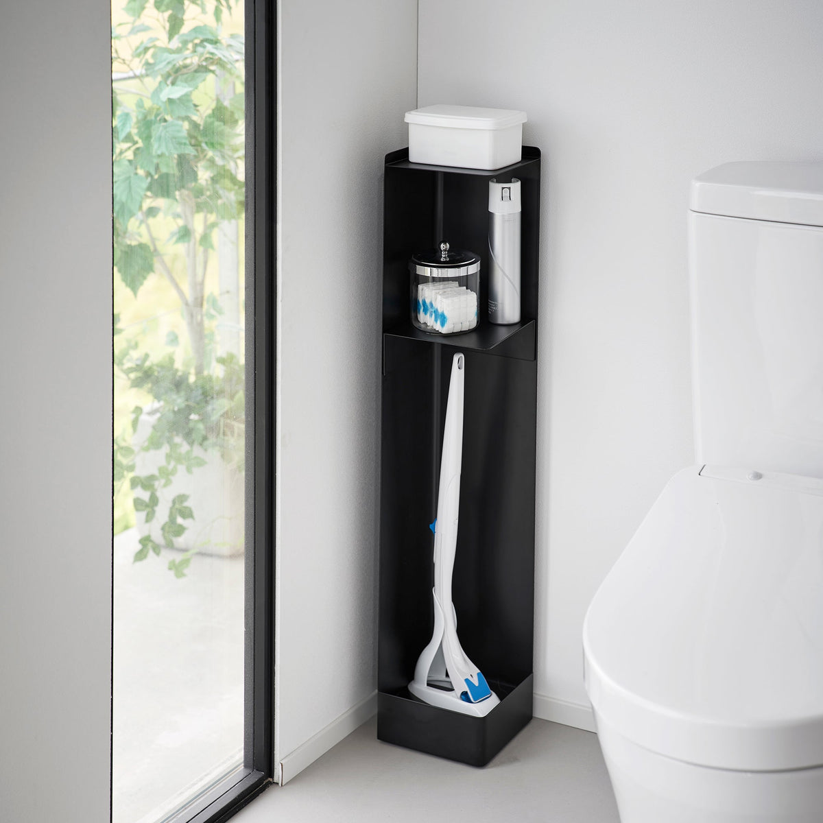 Black Yamazaki Home Rolling Bathroom Organizer - Steel beside a white toilet and window, holding a white tissue box, small bathroom supplies and long white cleaning tools.