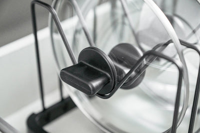 Close-up of the black Yamazaki Home Adjustable Pot Lid Organizer - Steel base rail, showing repeated rectangular slots and upright divider wires beneath stored lids.