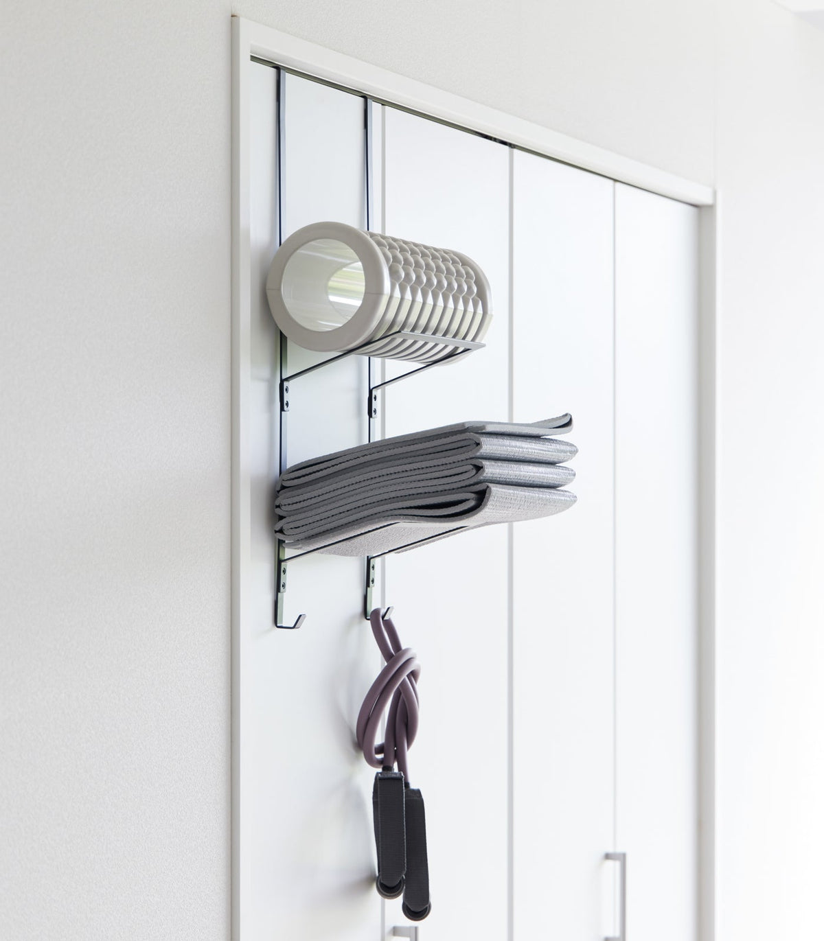 Black Yamazaki Home Over-the-Door Fitness Equipment Hanger - Steel on a white sliding closet door, with a grey foam roller on top, folded mat below and resistance bands hanging underneath.