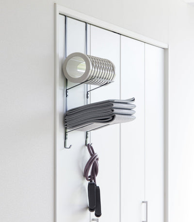 Black Yamazaki Home Over-the-Door Fitness Equipment Hanger - Steel on a white sliding closet door, with a grey foam roller on top, folded mat below and resistance bands hanging underneath.
