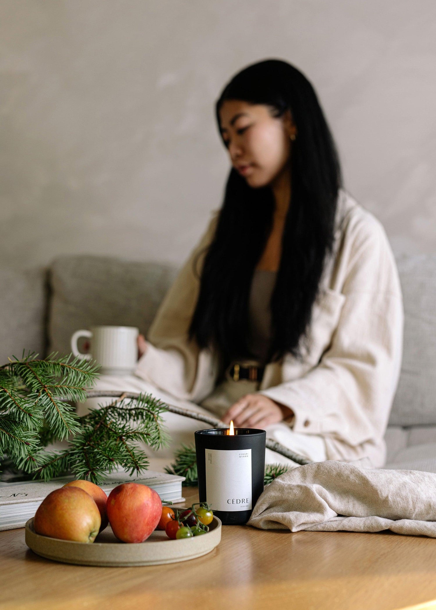Woman sitting at a table with a candle, fruit, and greenery in a cozy indoor setting.