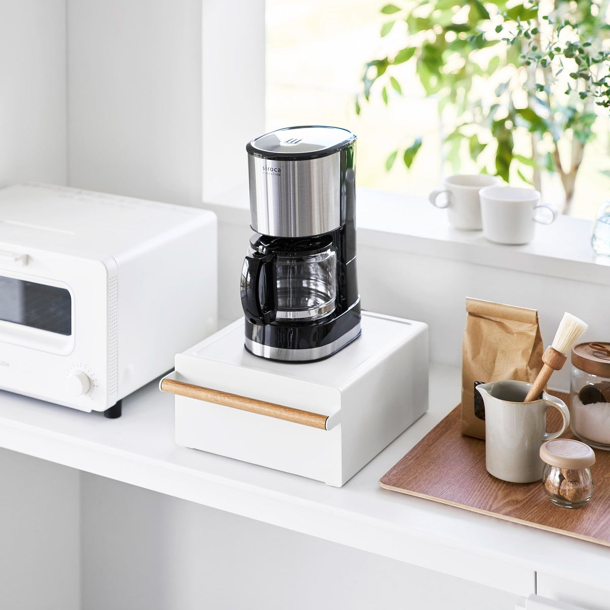 White Yamazaki Home Countertop Rack with Drawer - Steel + Wood supporting a black-and-silver drip coffee maker beside an oven, mugs and coffee supplies.