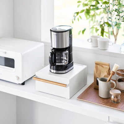White Yamazaki Home Countertop Rack with Drawer - Steel + Wood supporting a black-and-silver drip coffee maker beside an oven, mugs and coffee supplies.