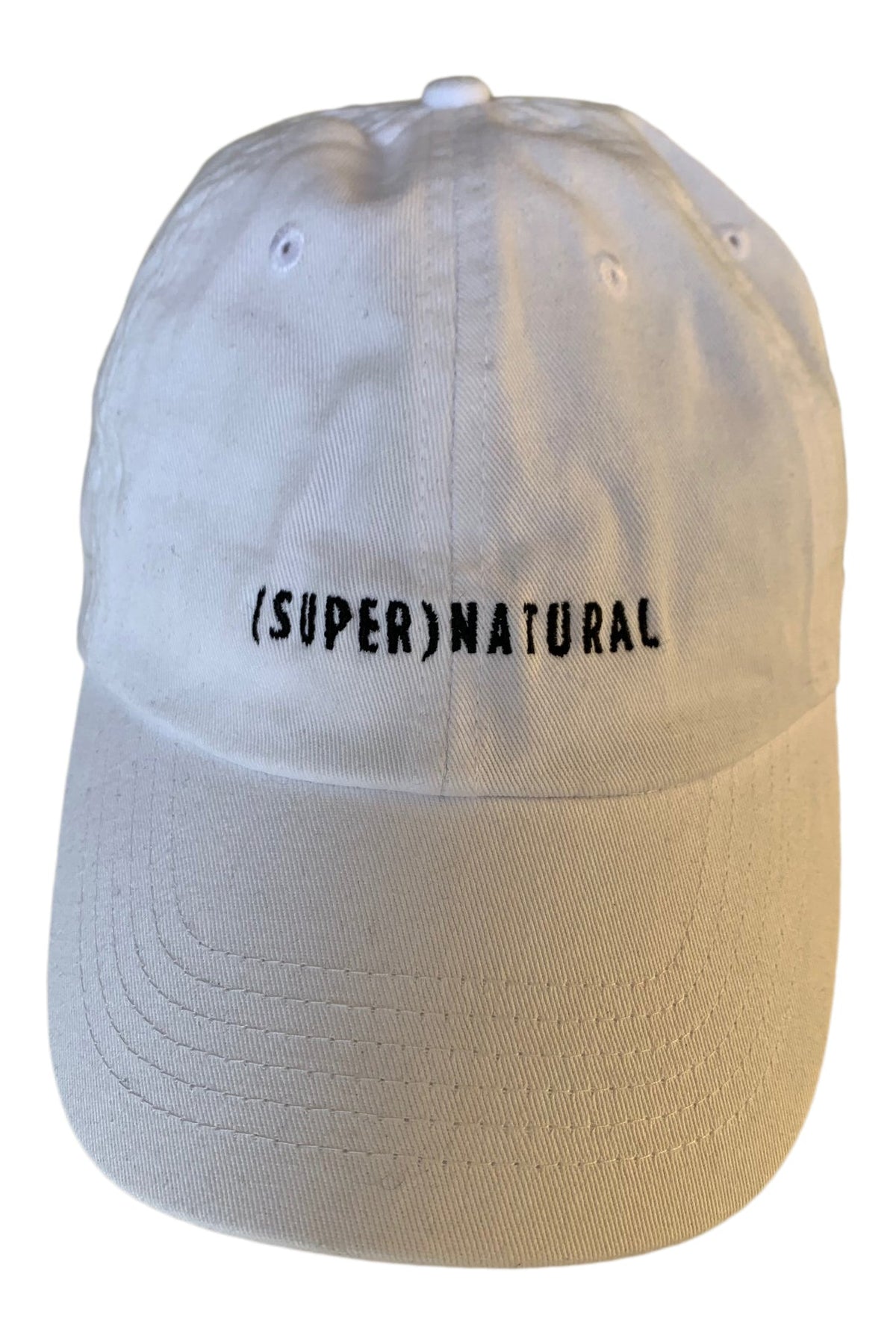 Front three-quarter view of a cream six-panel baseball cap with a curved brim and black embroidered “(SUPER)NATURAL” lettering.