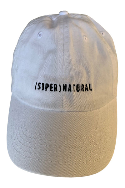 Front three-quarter view of a cream six-panel baseball cap with a curved brim and black embroidered “(SUPER)NATURAL” lettering.