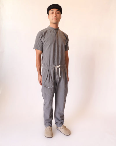 Model wearing a gray short-sleeve jumpsuit with a band collar, front closure, drawstring waist, and utility pockets.