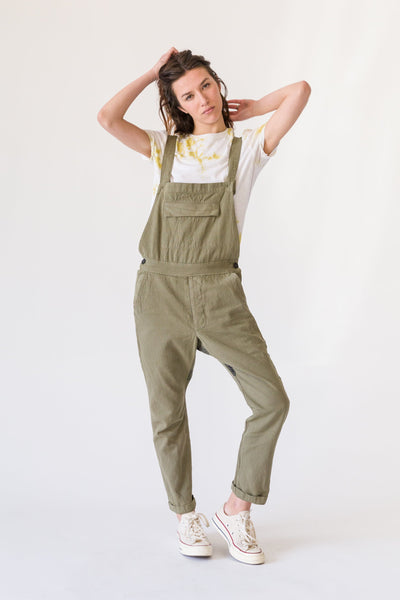 Model posing in olive-green overalls over a white and yellow printed T-shirt with white sneakers.