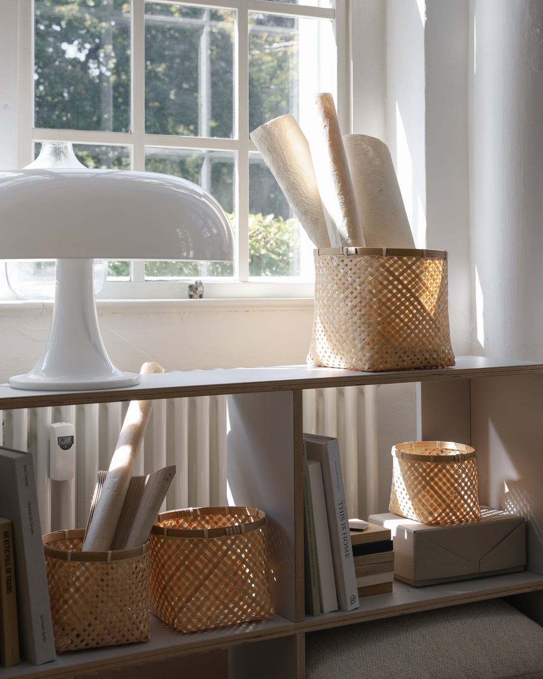 Bamboo baskets organizing rolled paper, art supplies, books, and small objects on a sunlit shelf.