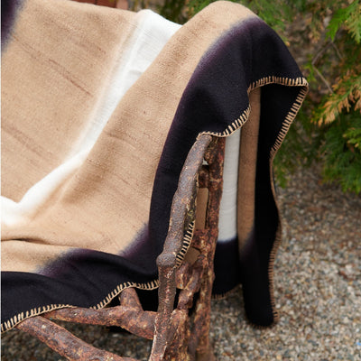 Siro Brown throw draped over an outdoor wood chair, highlighting its soft tan weave, black border and hand stitching