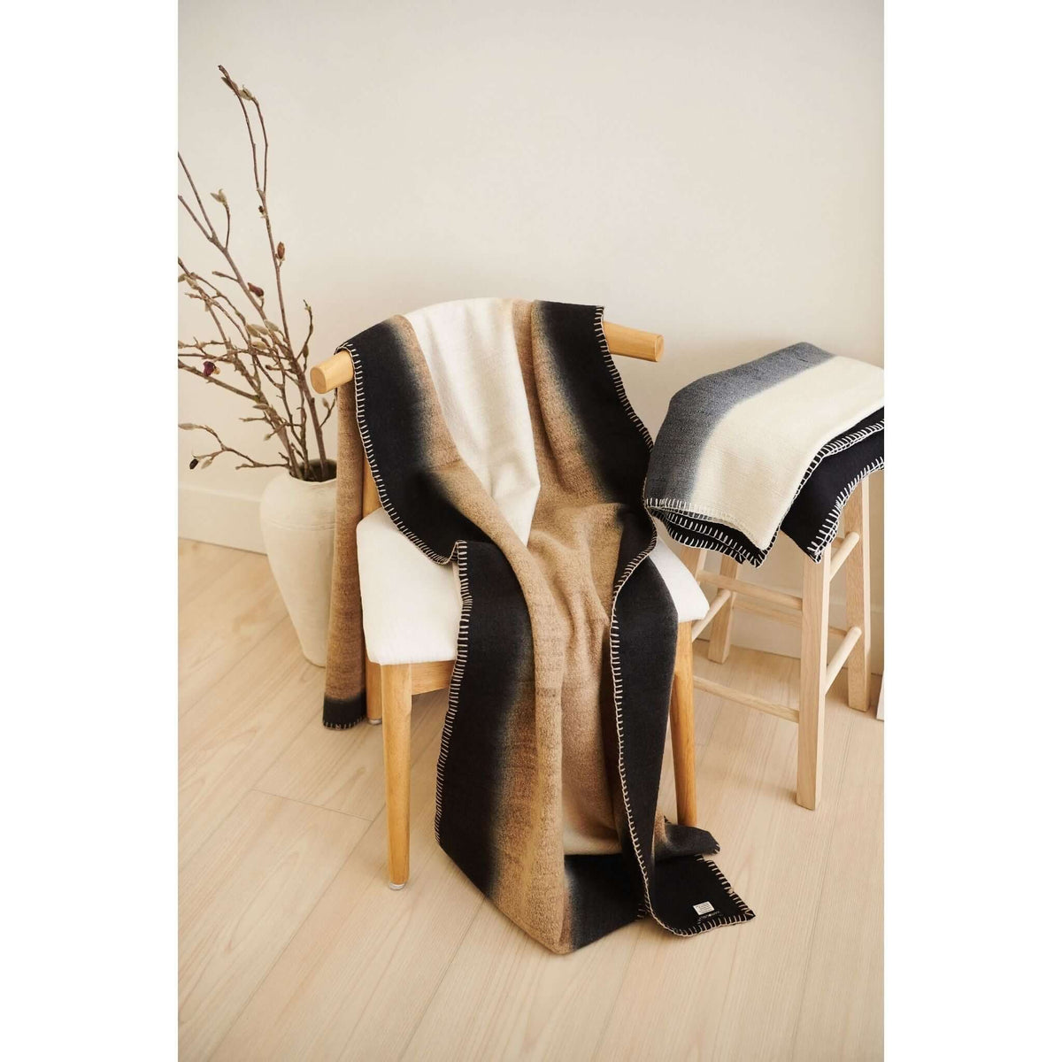 Siro Brown merino throw folded over a white sculptural stool beside smooth stones and a natural tree branch