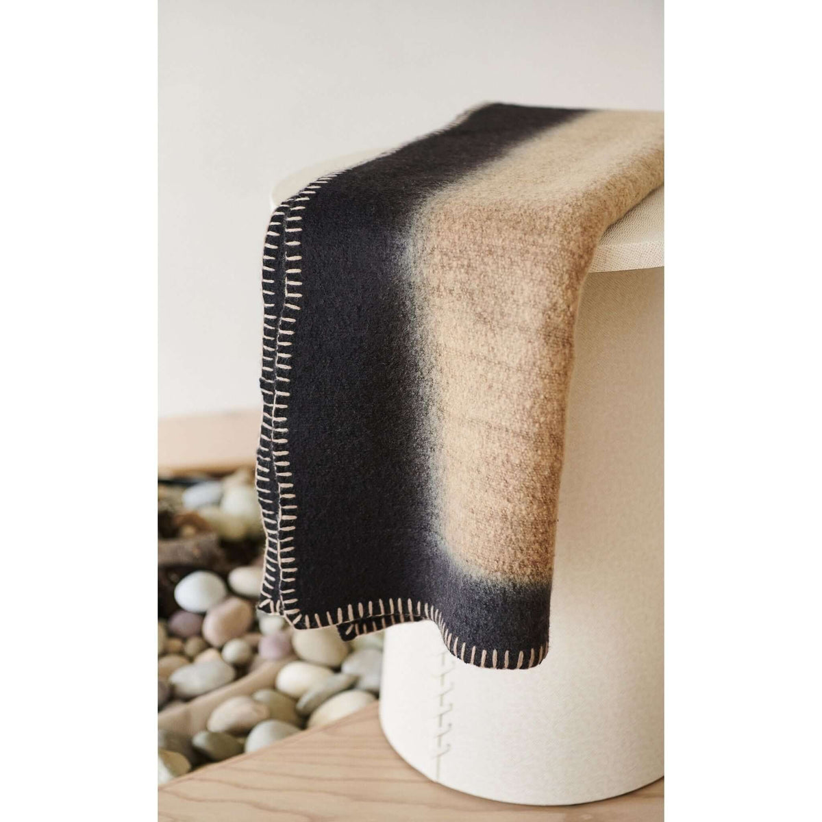 Siro Brown handwoven throw displayed on a white cylindrical stool in a natural setting with stones and wood