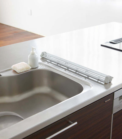 Yamazaki Home Over-the-Sink Dish Drainer - Steel rolled into a compact bundle along the rear edge of a sink, beside a white dispenser and sponge.