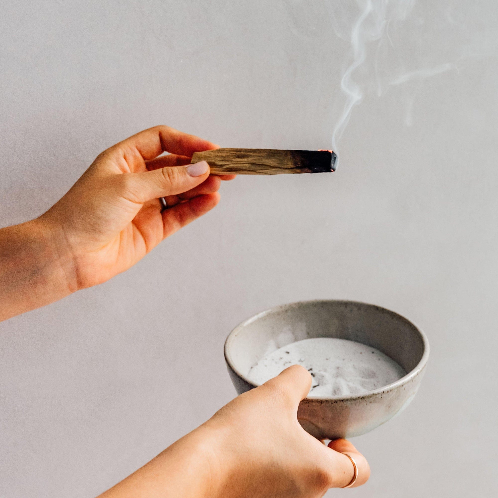 Hand holding a lit incense stick above a bowl of white powder on a neutral background