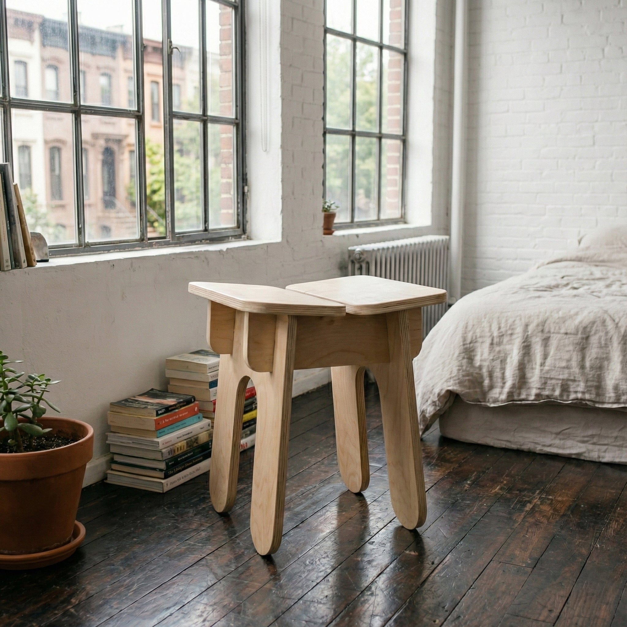 BURIJ natural wood stool beside a bedroom window with stacked books, a potted plant and an unmade bed
