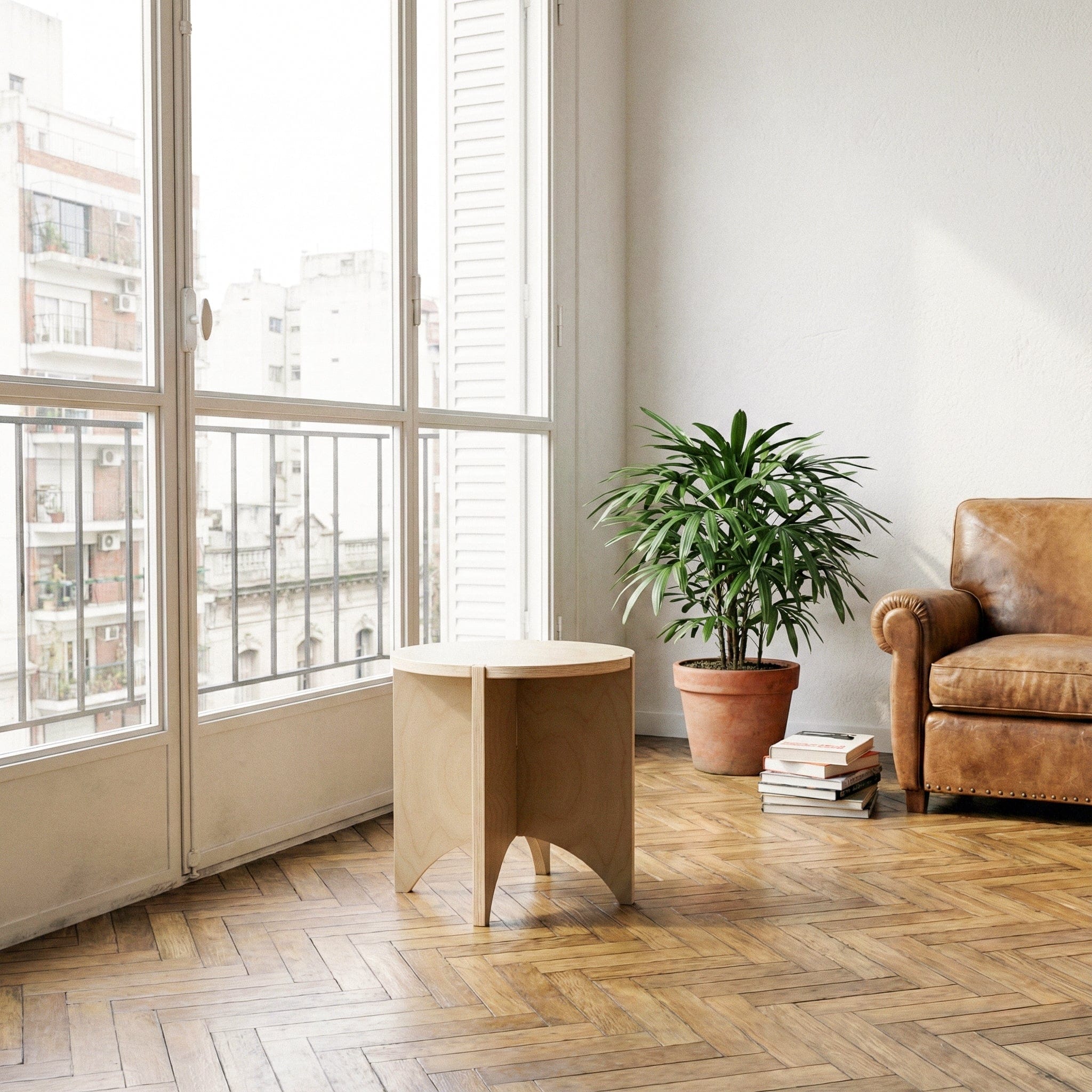 YOYO round wood end table in a sunlit living room with tall windows, a potted plant, books and a brown leather chair