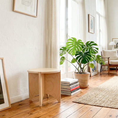 YOYO wood end table against a sunlit wall beside stacked books, a large leafy plant and a woven rug