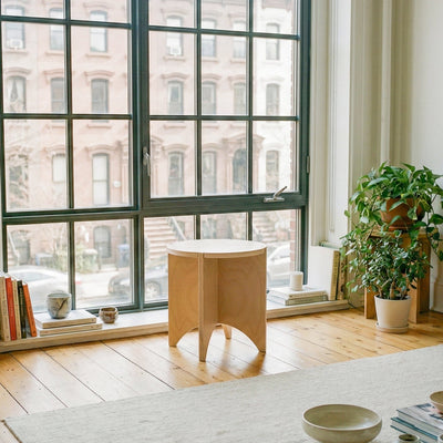YOYO round wood end table positioned in front of tall city windows beside potted plants and books