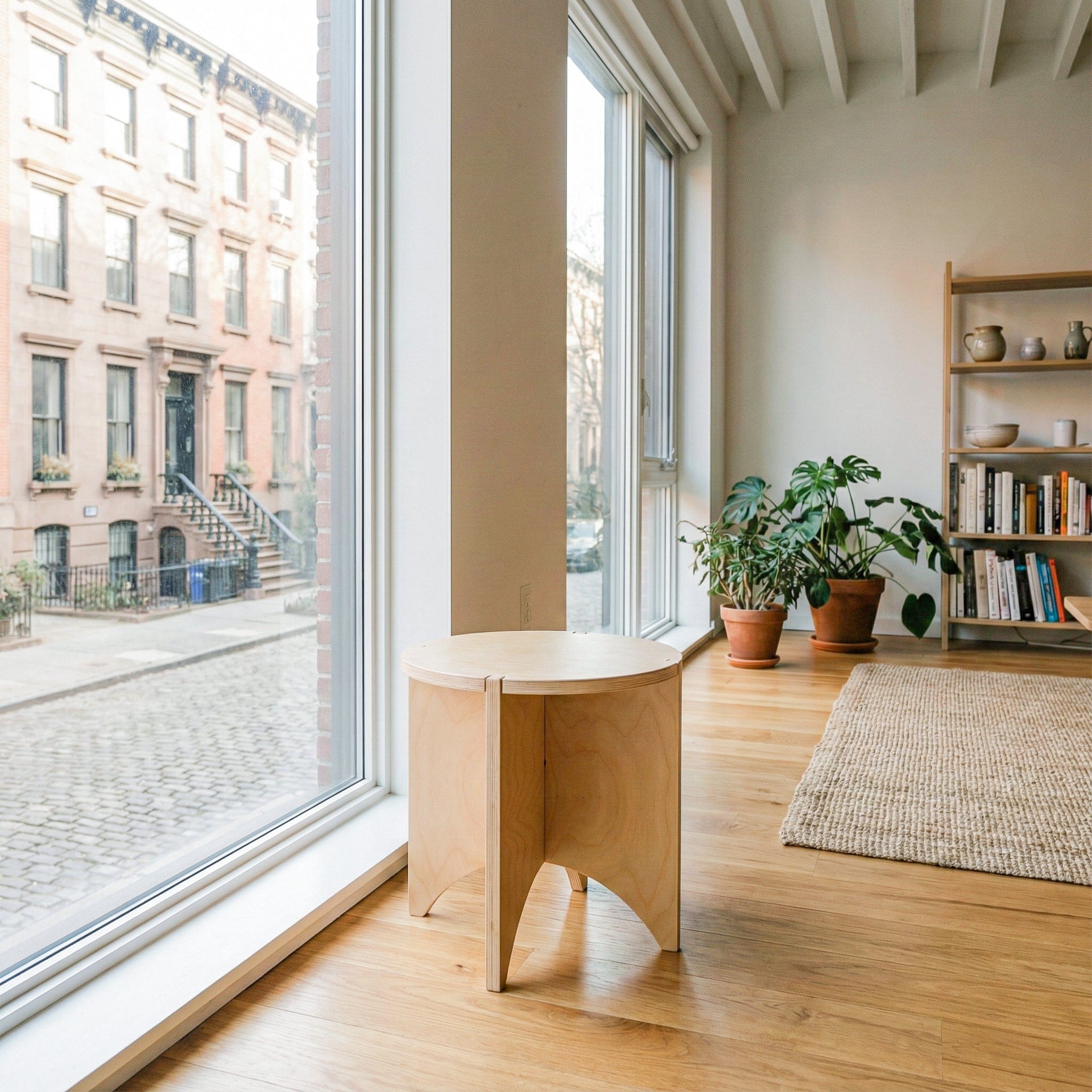 YOYO natural wood end table beside a floor-to-ceiling window in a bright apartment with wood floors, plants and bookshelves