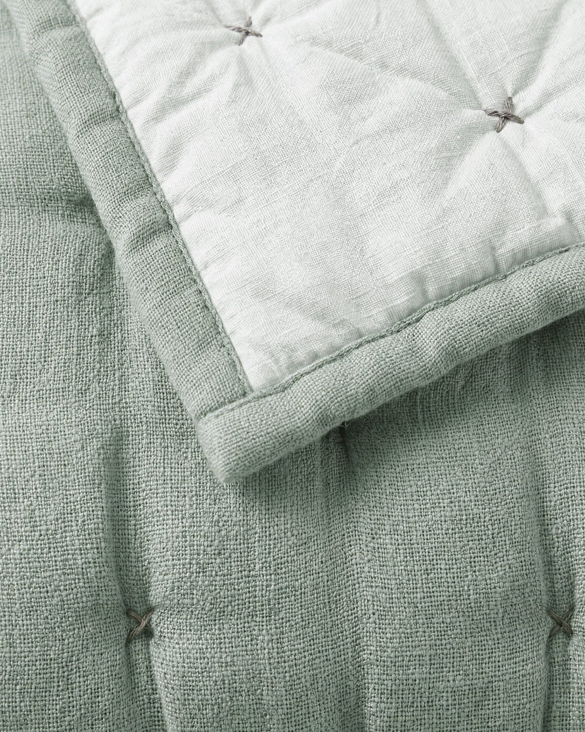 Close-up of the sage quilt’s textured surface and tufted stitches, with the pale reverse folded over a matching green border.