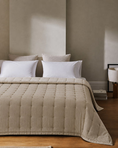 Taupe quilt spread across a bed with white pillows, draping over the sides and foot in a neutral bedroom.
