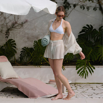 Model walking barefoot across a patio in the open cover-up over a white bikini top and striped shorts, carrying a rolled green towel beside tropical plants.