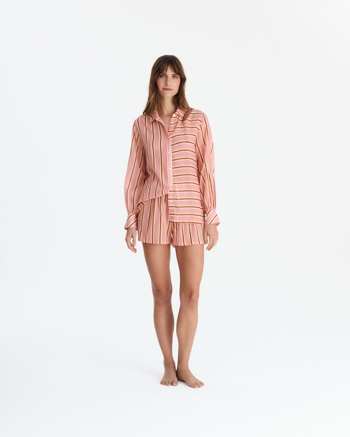 Front view of a barefoot model wearing coral pink striped pajamas, with a loose long-sleeve button-front shirt featuring contrasting stripe directions and matching vertically striped shorts.