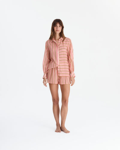 Front view of a barefoot model wearing coral pink striped pajamas, with a loose long-sleeve button-front shirt featuring contrasting stripe directions and matching vertically striped shorts.