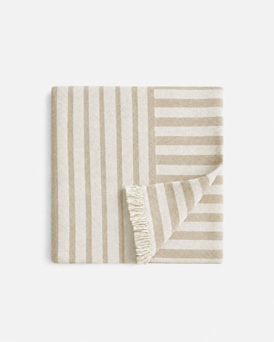 Folded beige and cream Serena Beach Blanket with contrasting vertical and horizontal stripes, with one corner turned back to reveal cream fringe.