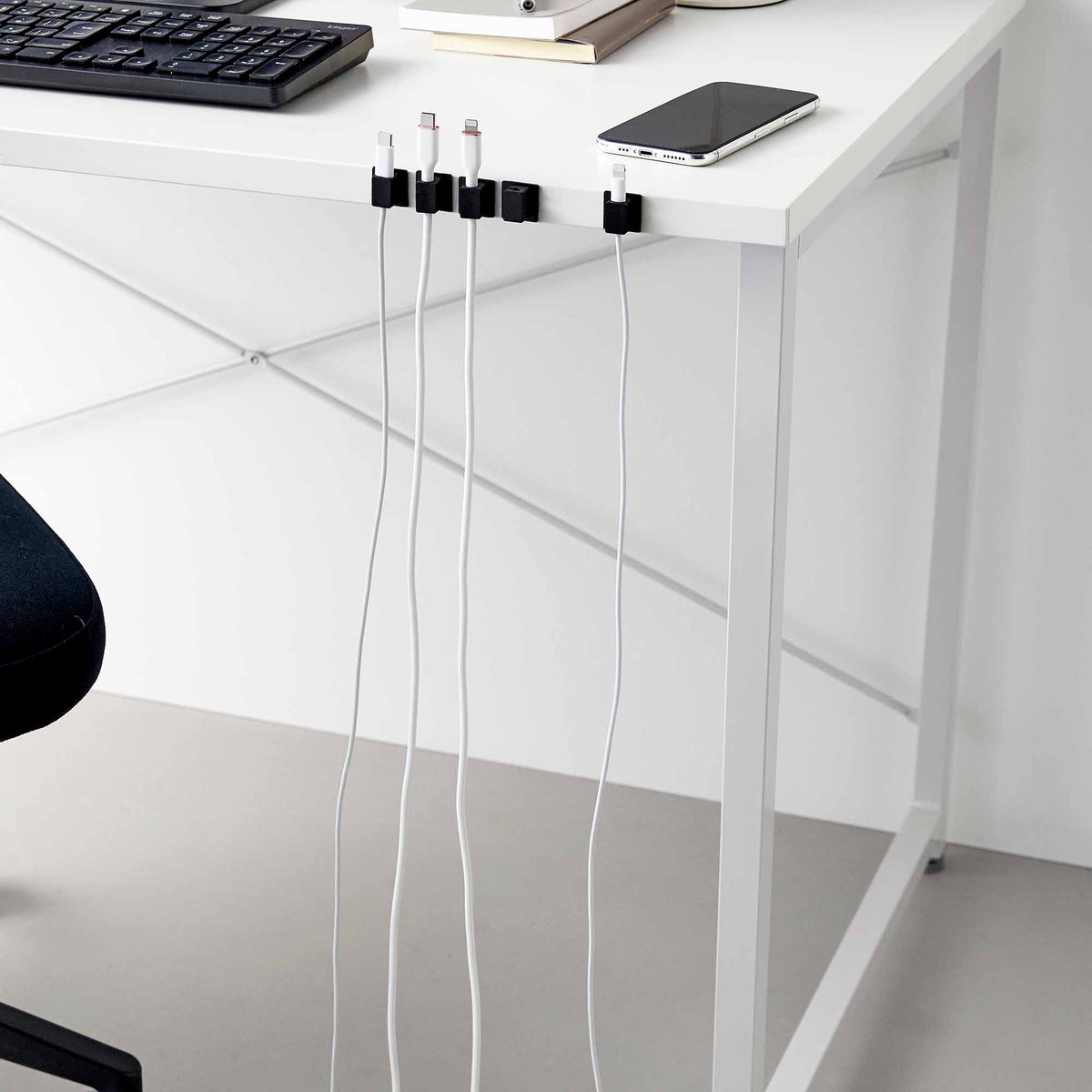 Wide view of black Yamazaki Home Magnetic Cable Holders With Adhesive Steel Bases (Set of 5) - Silicone attached along a white desk edge, with white cables hanging individually below the keyboard and phone.