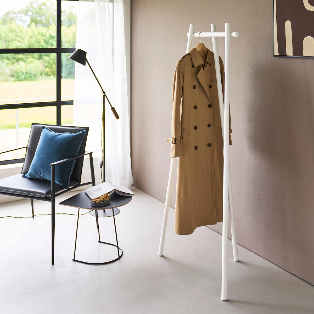 White Yamazaki Home Tri-Post Coat Rack (60" H) - Steel holding a single tan trench coat on a wooden hanger, showing the long garment suspended between its angled legs in a bright room.