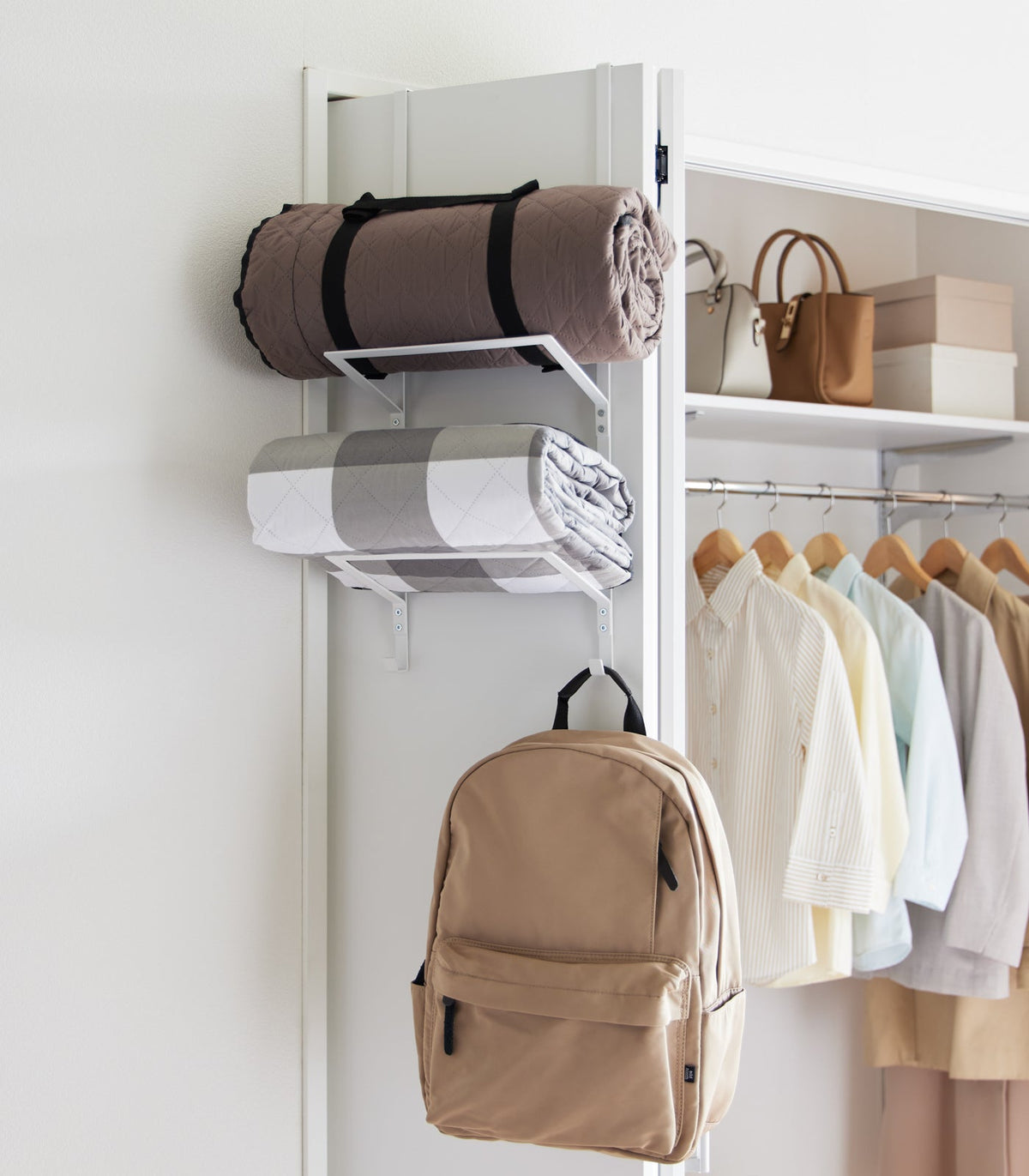 White Yamazaki Home Over-the-Door Fitness Equipment Hanger - Steel holding a rolled brown blanket, folded grey-and-white textile and tan backpack beside an open wardrobe.