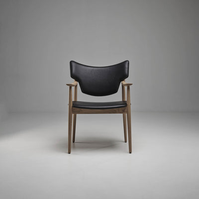 Front view of the black Eikund Veng Arm Chair in a gray studio, showing its curved upholstered back, separate seat, and slender brown wood frame.