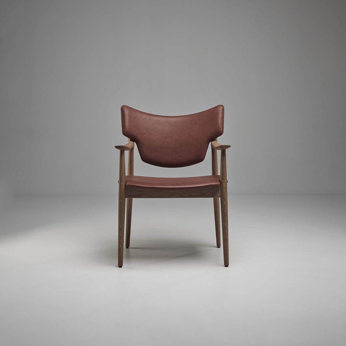 Front view of the reddish-brown Eikund Veng Arm Chair in a gray studio, showing its wing-shaped upholstered back, matching seat, and dark brown wood frame.