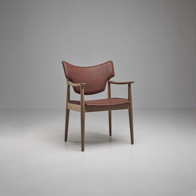 Three-quarter front view of the reddish-brown Eikund Veng Arm Chair, showing the curved back, slim wood armrests, and open space between the backrest and seat.