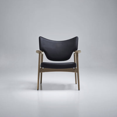 Front view of the Eikund Veng Lounge Chair with black upholstery and a light wood frame in a gray studio, showing the curved back, separate seat cushion, and tapered legs.