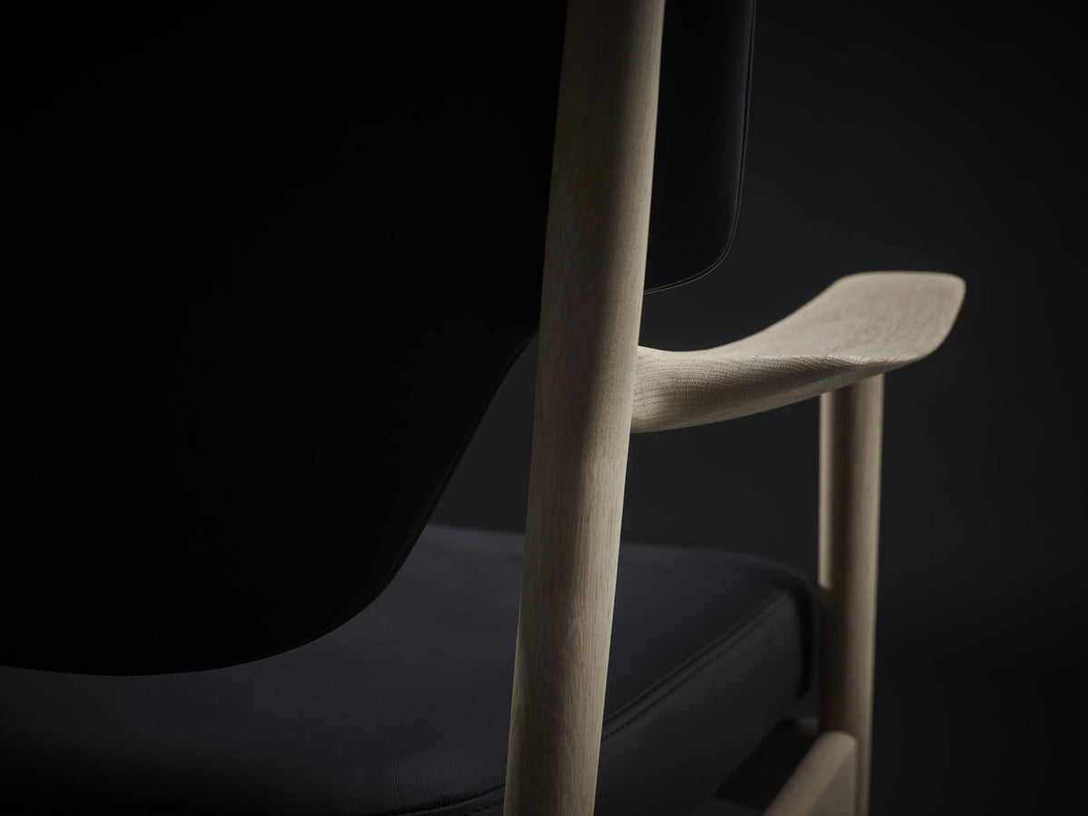 Side detail of the Eikund Veng Lounge Chair, showing a slender wood upright, the upturned armrest tip, and the rounded edge of the black seat cushion.