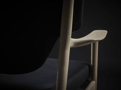 Side detail of the Eikund Veng Lounge Chair, showing a slender wood upright, the upturned armrest tip, and the rounded edge of the black seat cushion.