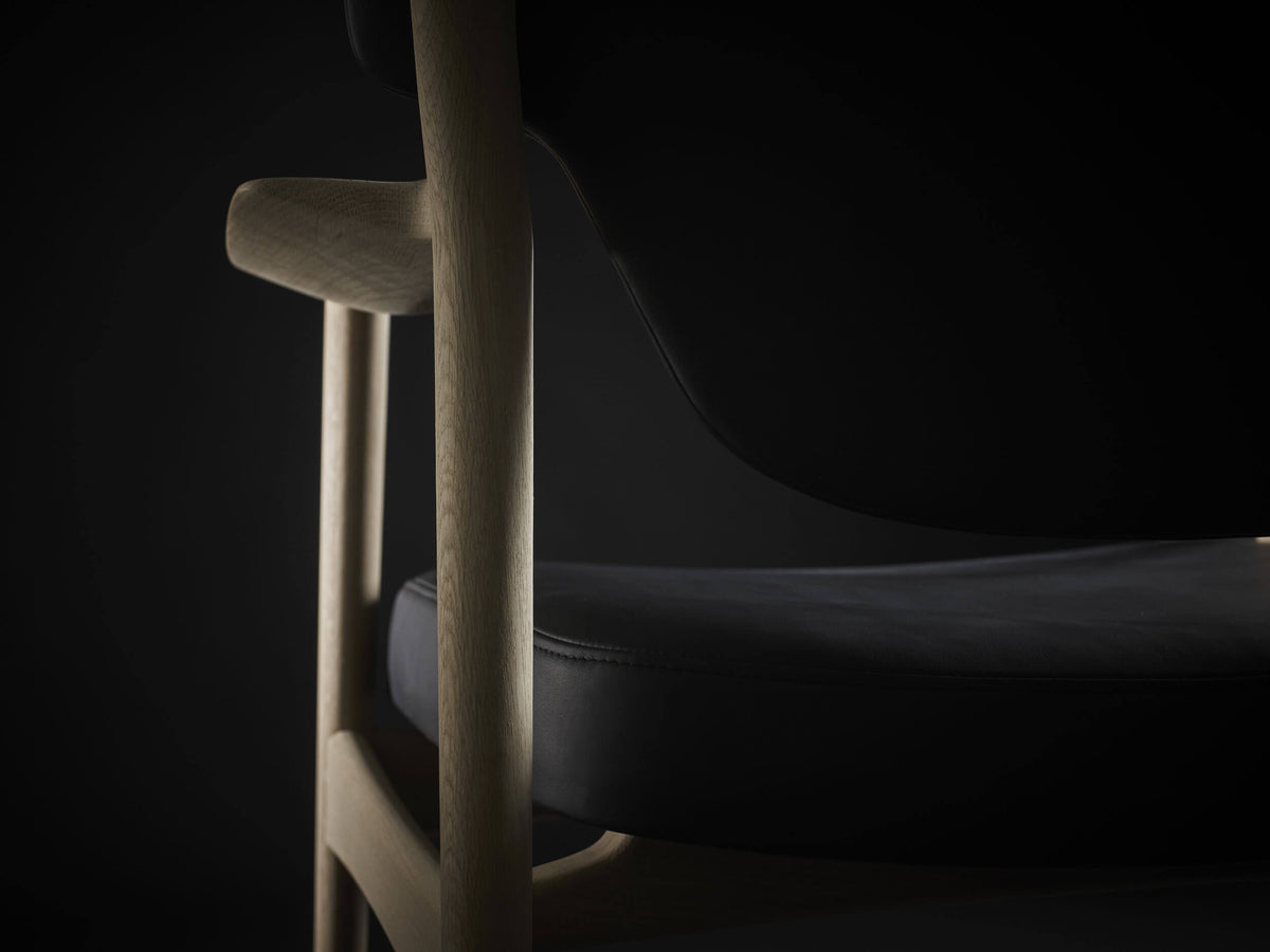 Close-up through the Eikund Veng Lounge Chair’s light wood side frame, showing the armrest supports, curved lower backrest, and black padded seat.
