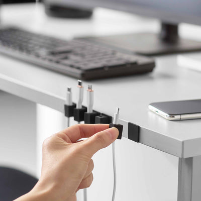A hand removes a black Yamazaki Home Magnetic Cable Holder With Adhesive Steel Base - Silicone from the desk edge, showing its placement beside other holders and charging cables.