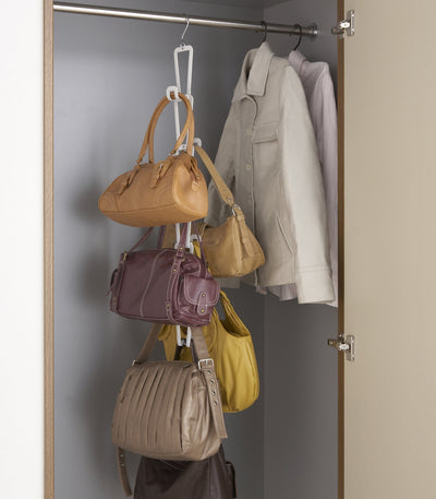 White Yamazaki Home Cascading Storage Hooks - Steel suspended from a closet rail, storing several tan, purple, yellow and grey handbags vertically beside pale coats.