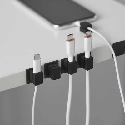 Black Yamazaki Home Magnetic Cable Holders With Adhesive Steel Bases (Set of 5) - Silicone holding white charging cables upright along a pale desk edge beside a black phone.