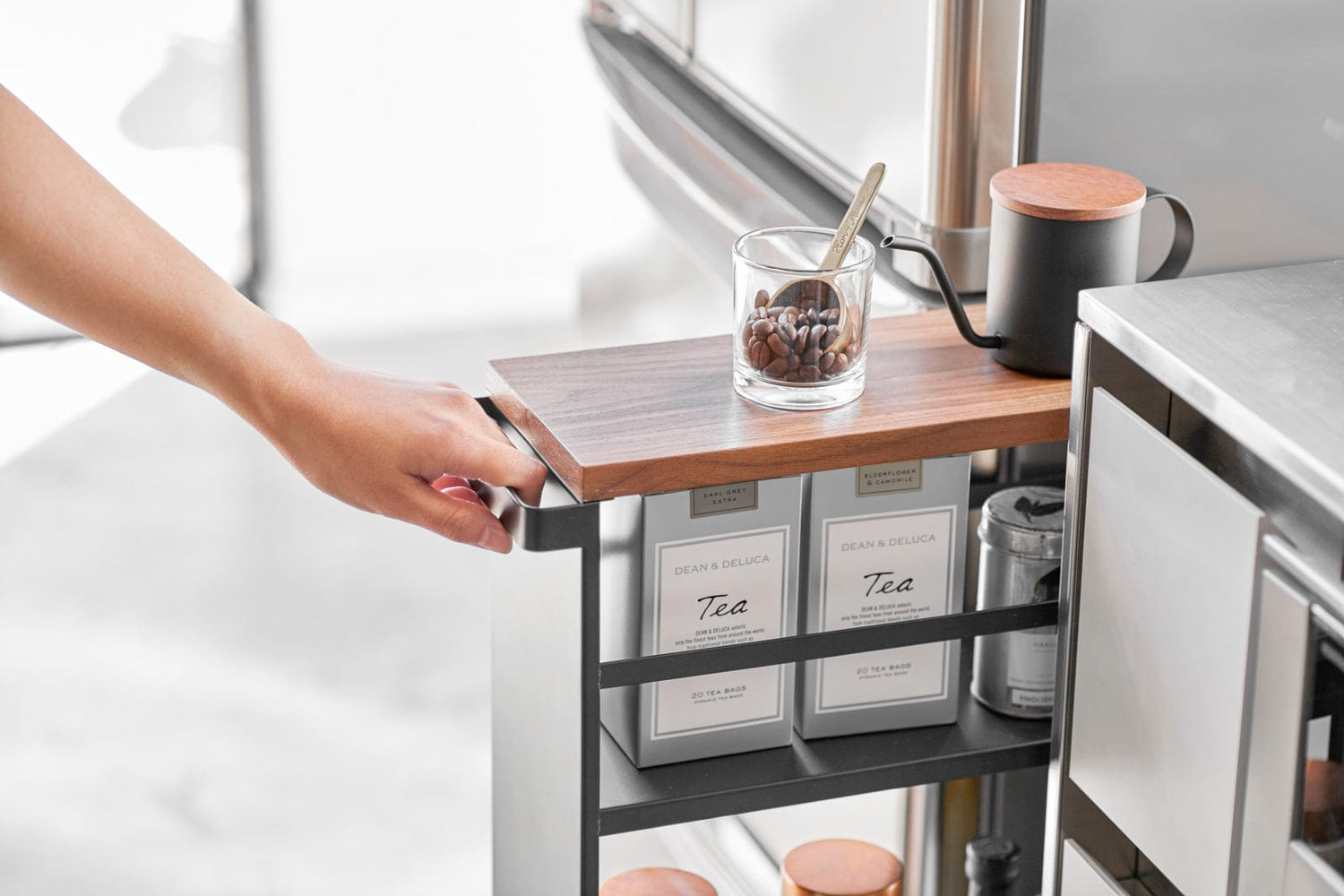 Kitchen cart with a wooden top holding a glass of coffee beans and a mug, next to tea bags.