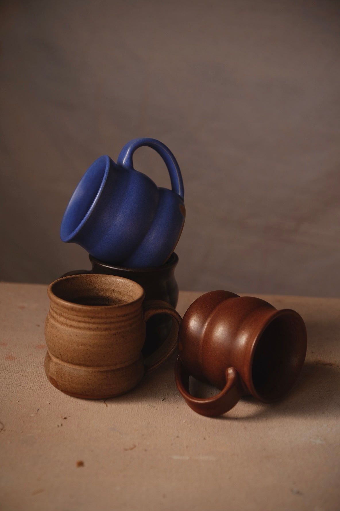 Blue, golden-brown and reddish-brown Settle Ceramics The Wave Mugs arranged on a brown tabletop, with one tilted above the others and another resting on its side, showing rounded ceramic bodies, broad horizontal contours and curved handles.