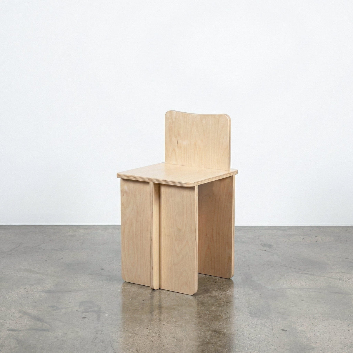 Studio Furnis Williamsburg natural wood chair with a gently curved backrest, flat square seat and broad panel legs on a polished concrete floor