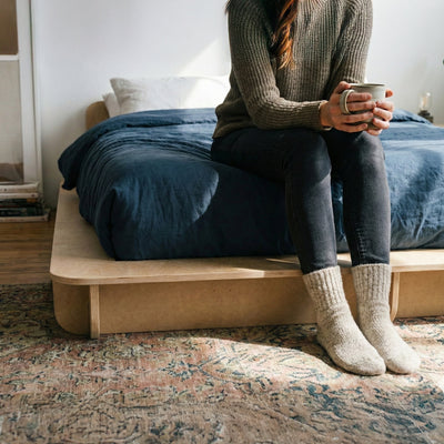 Person holding a mug while seated on the foot of a Studio Furnis bed platform with blue bedding