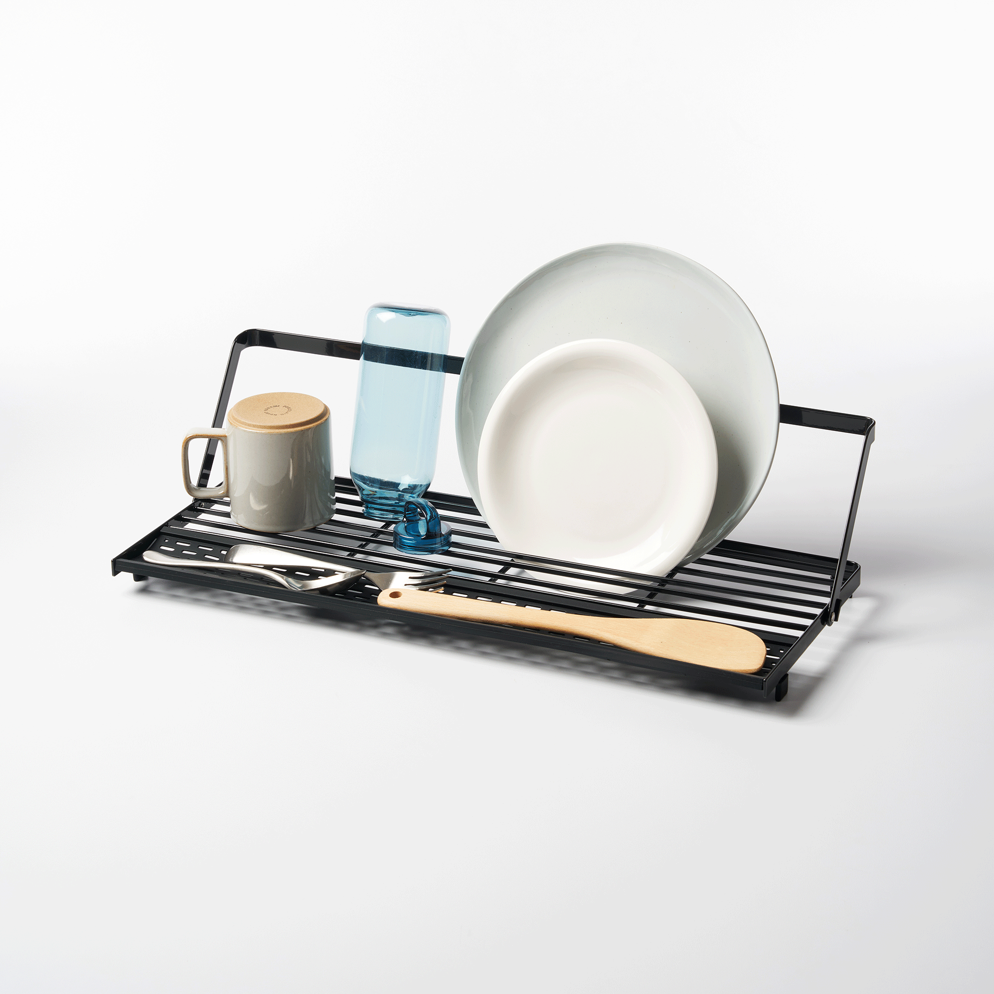 Angled view of the black Yamazaki Home Slim Over-the-Sink Dish Drying Rack - Steel loaded with upright plates, a blue bottle and mug, with utensils resting across the front bars.