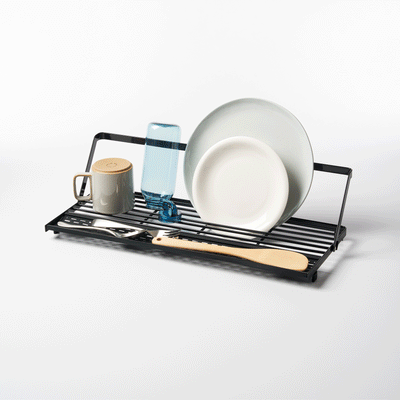 Angled view of the black Yamazaki Home Slim Over-the-Sink Dish Drying Rack - Steel loaded with upright plates, a blue bottle and mug, with utensils resting across the front bars.