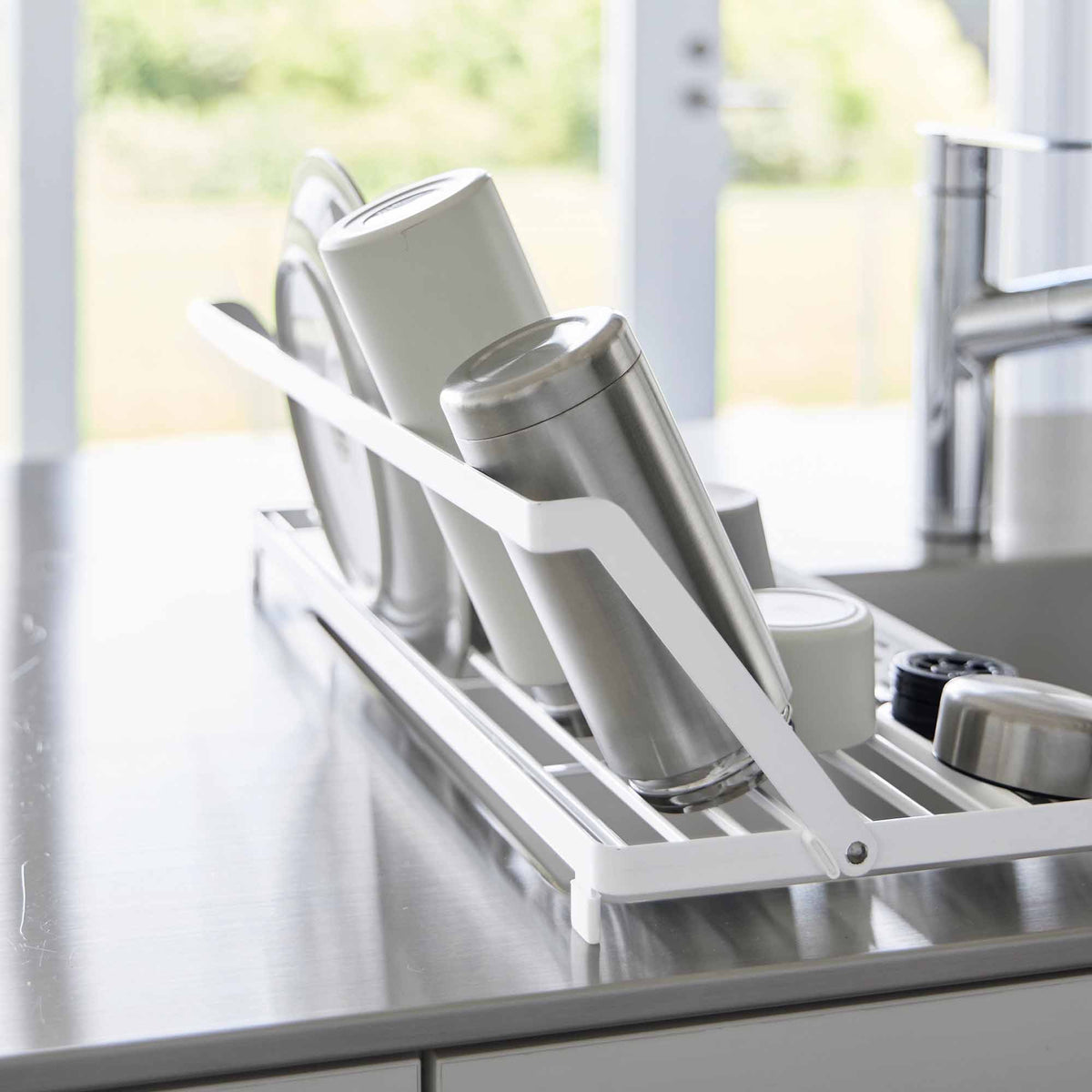 Side close-up of the white Yamazaki Home Slim Over-the-Sink Dish Drying Rack - Steel, showing tall cups and tumblers leaning against the raised rail above the sink.