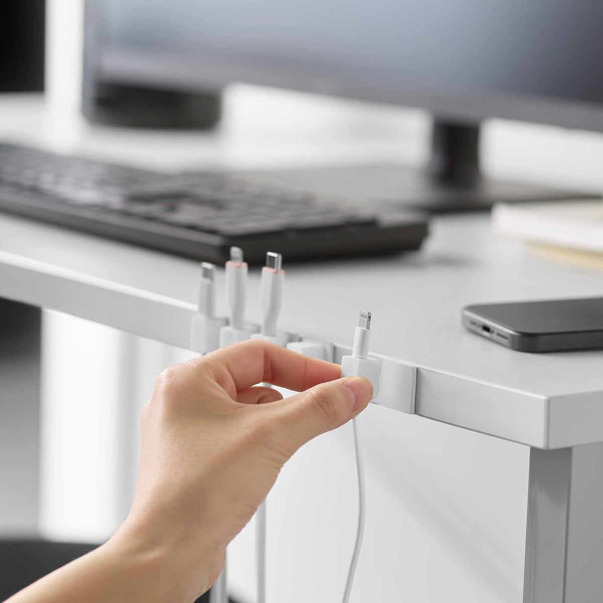 A hand lifts a white Yamazaki Home Magnetic Cable Holder With Adhesive Steel Base - Silicone from the desk edge while neighbouring holders keep charging connectors upright.