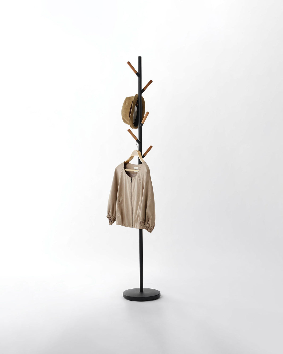 Black Yamazaki Home Coat Rack (70" H) - Steel holding a tan hat and beige jacket on a pale background, with warm brown wood tips contrasting against its dark pole.
